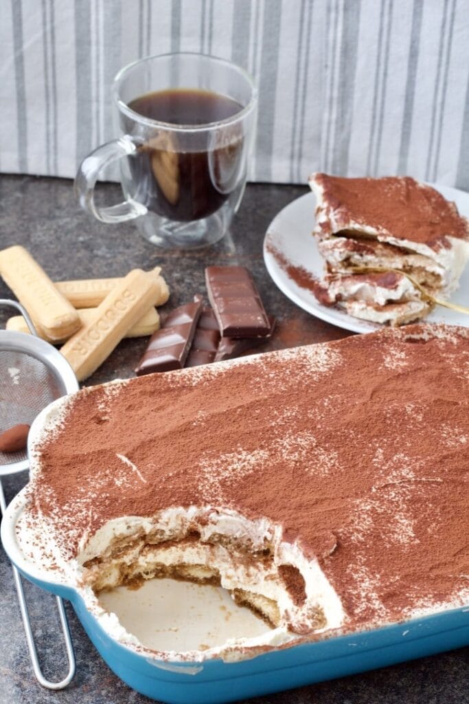 Easy Eggless Tiramisu - dish with tiramisu, one portion on a plate, cup of black coffee, chocolate pieces and lady fingers biscuits