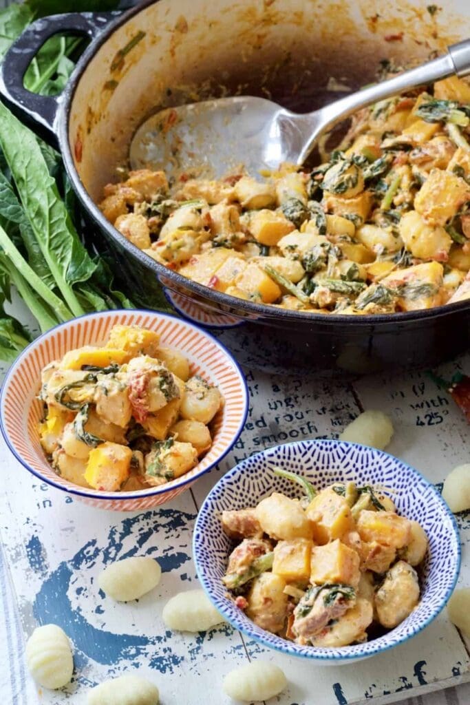 Easy Butternut Squash & Chorizo Gnocchi - two portions in small bowls with big pan in the background and uncooked gnocchi and fresh spinach styled around