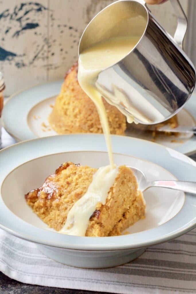 Custard being poured over steamed pudding slice.