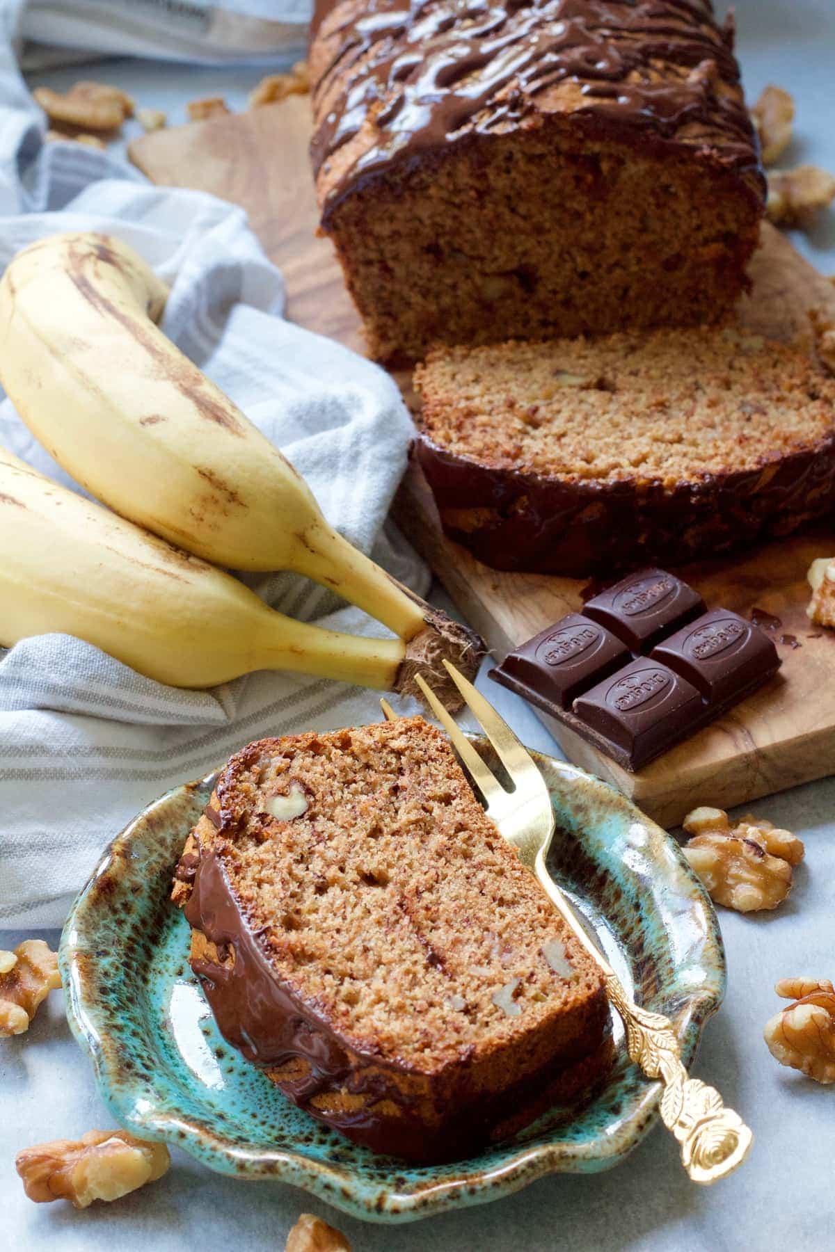 Banana loaf slice on a plate with loaf in the background.