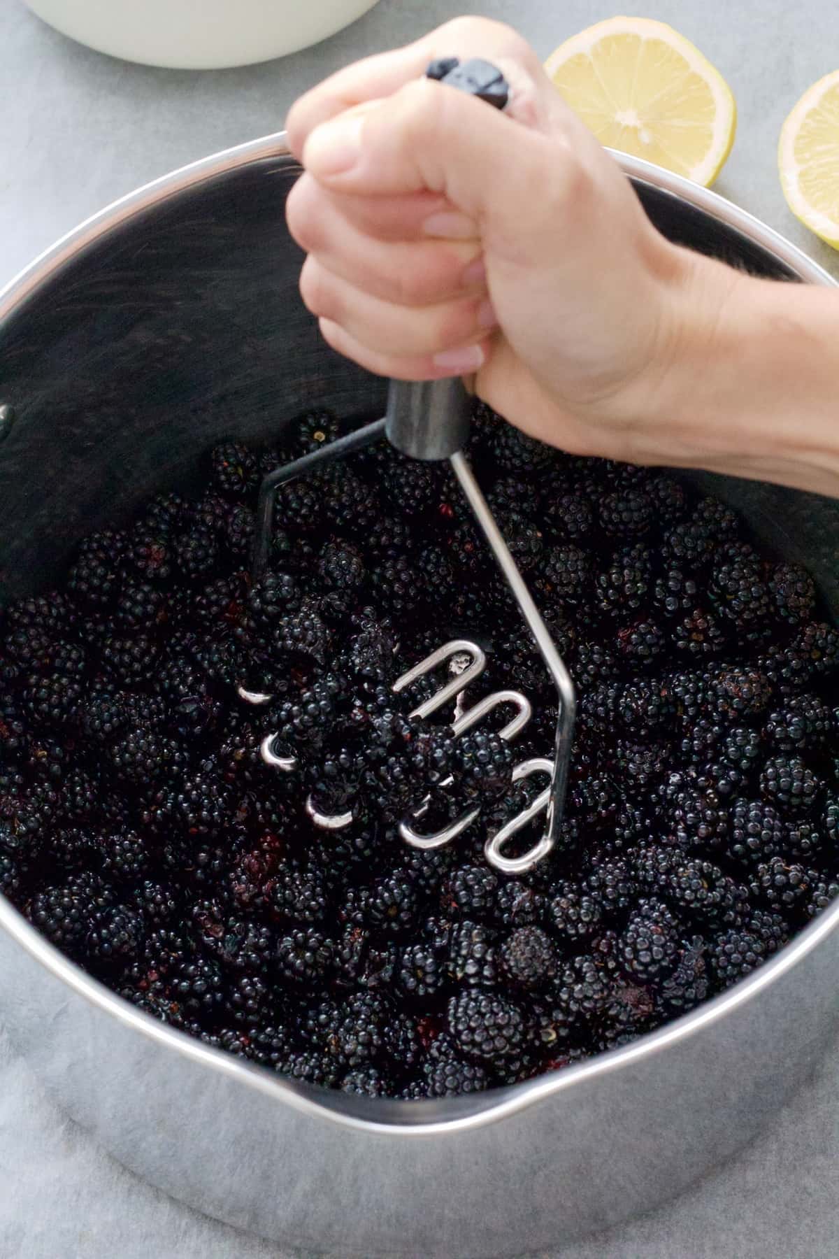 Hand mashing blackberries with potato masher in a pan.