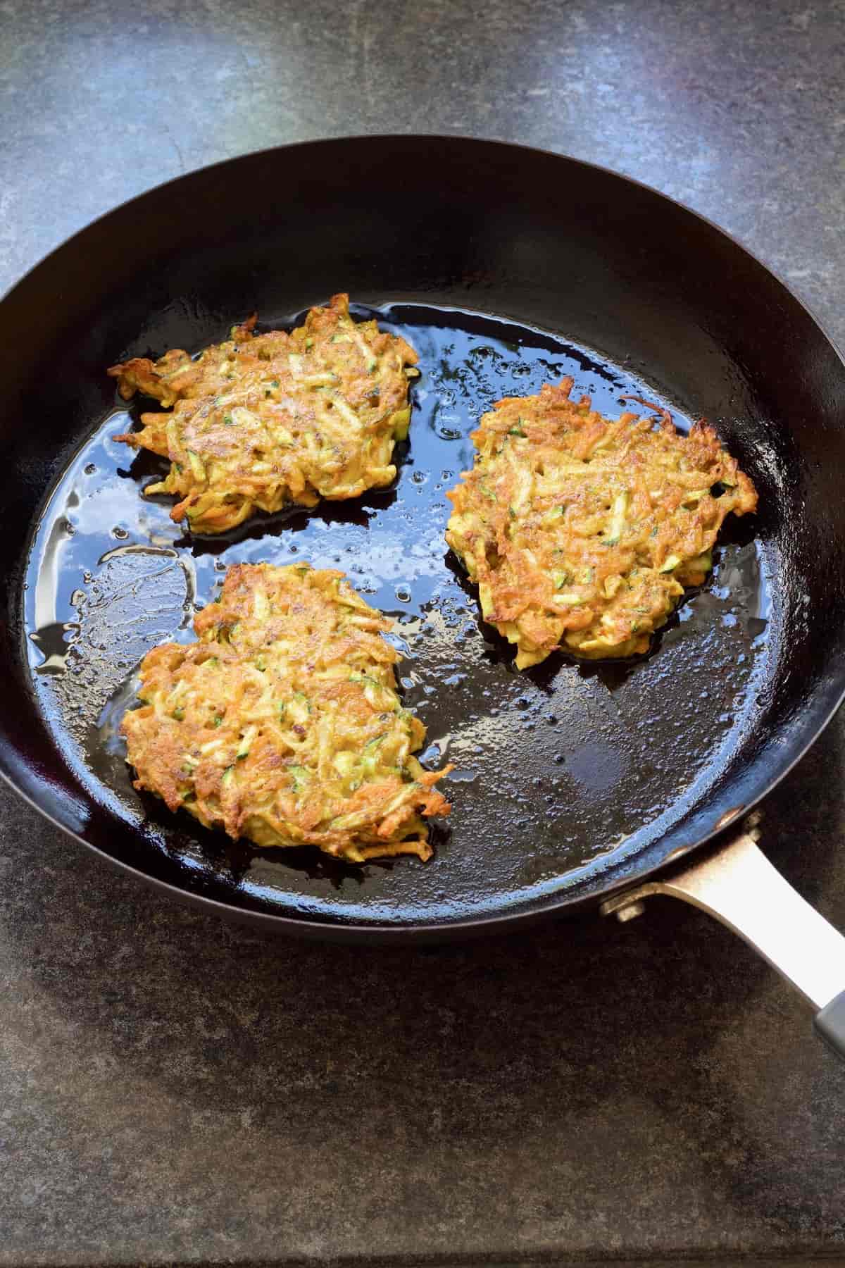 Three fritters in a frying pan, cooked side up.