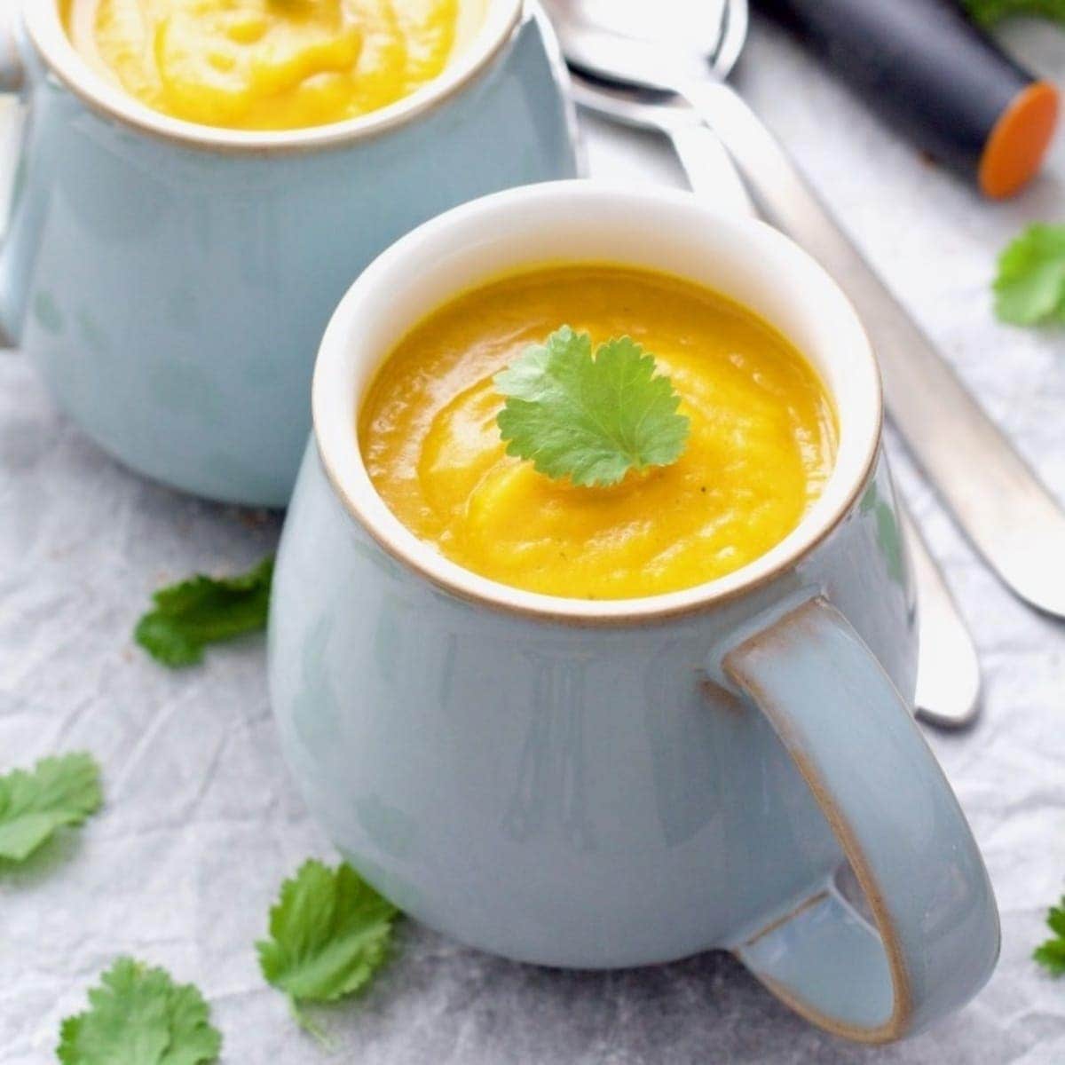 Mug filled with pumpkin soup garnished with coriander leaf.