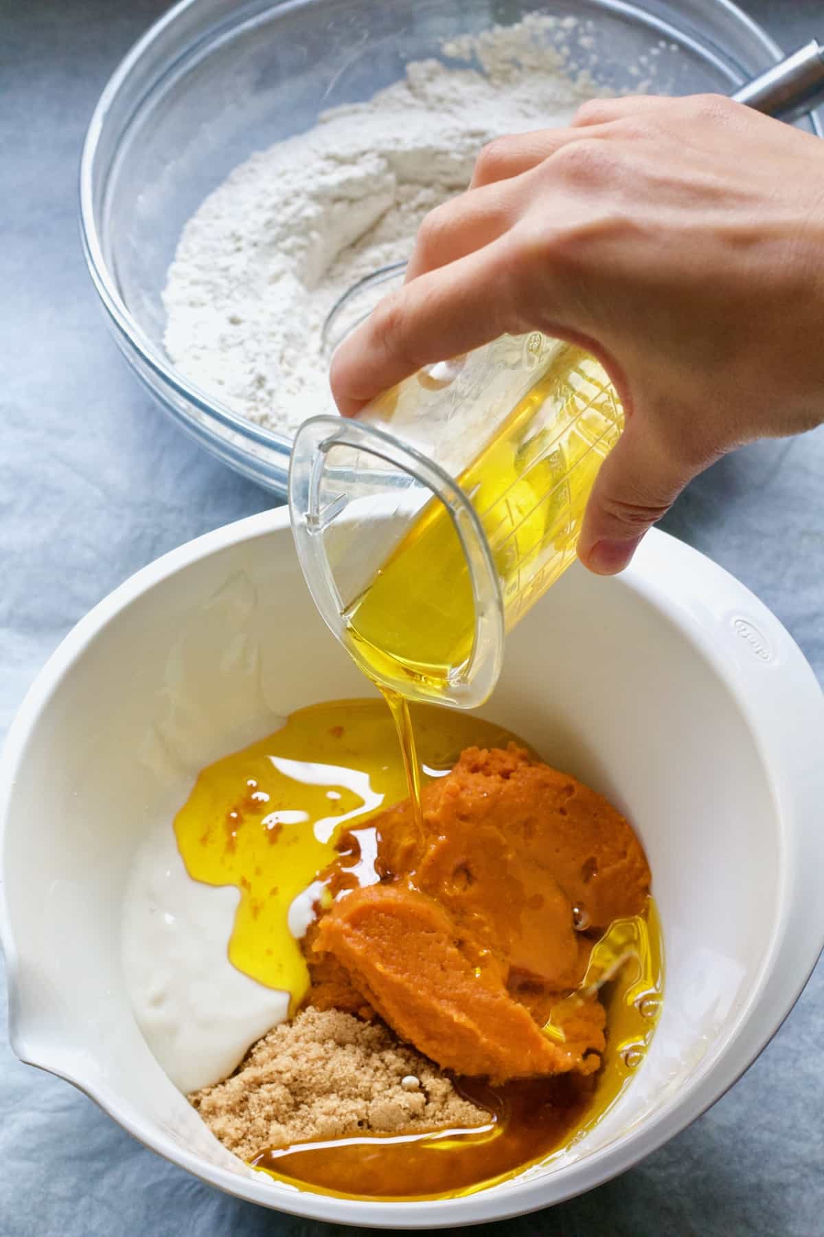 Hand pouring oil into yogurt, sugar and pumpkin puree.