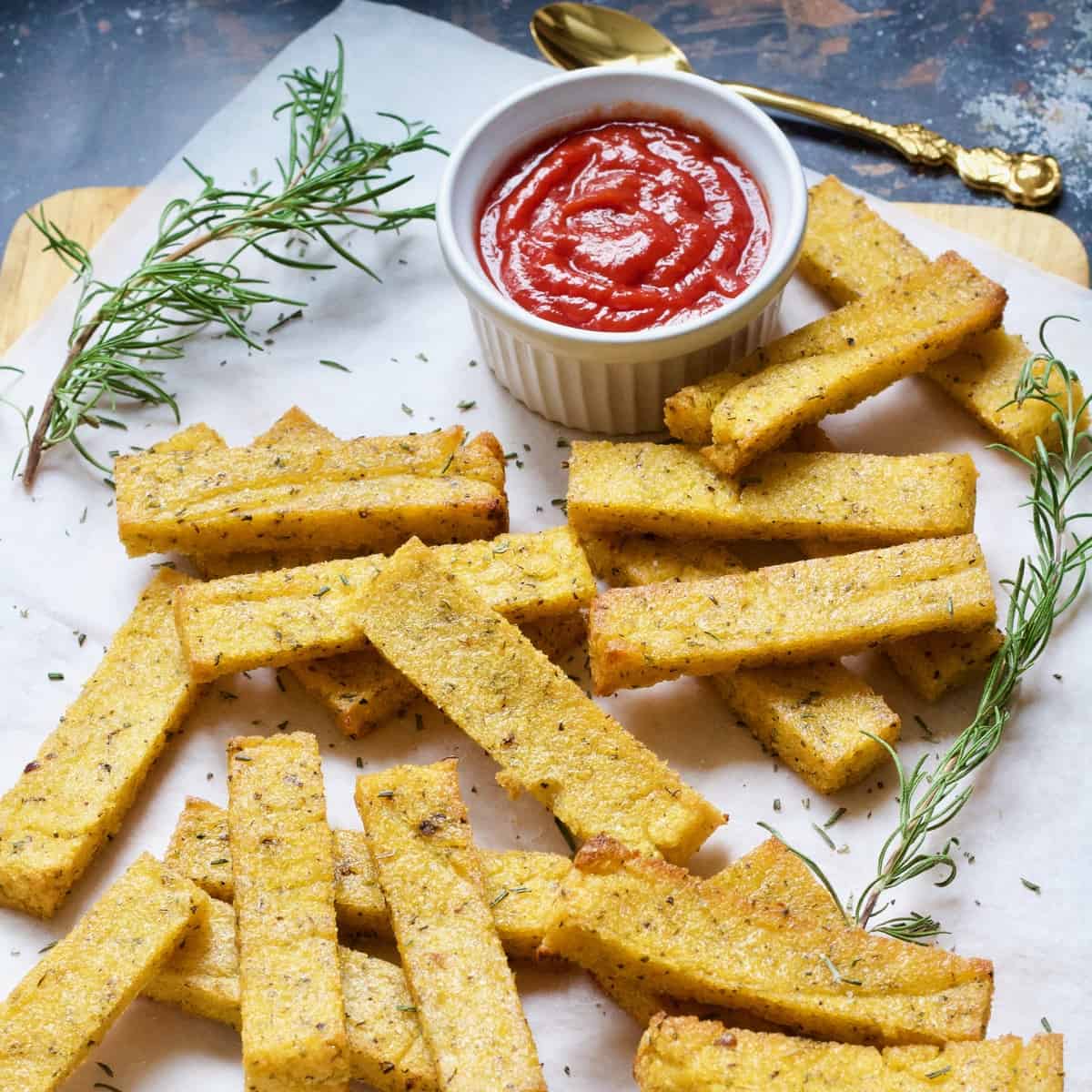 Pile of baked polenta chips with rosemary and bowl of ketchup.