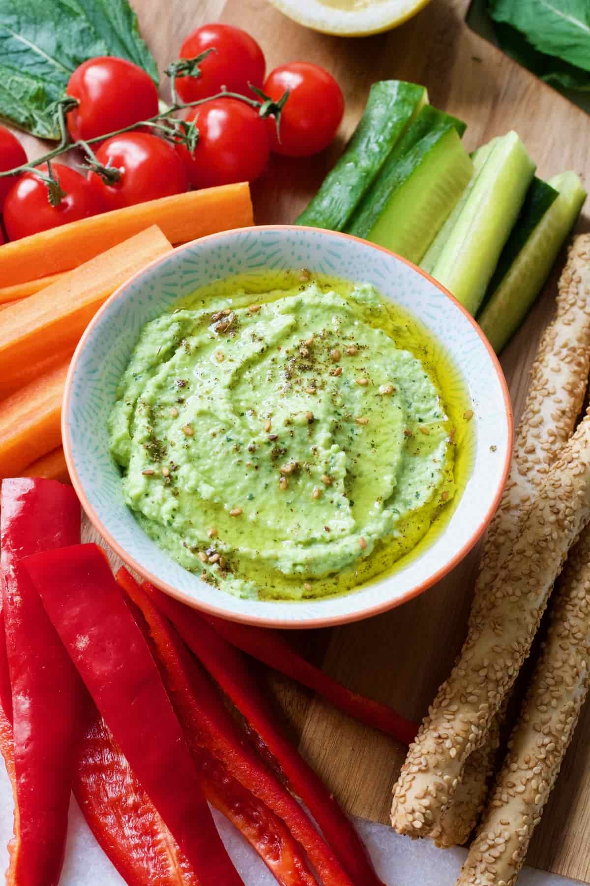 Broad bean dip in a bowl surrounded by veg & breadsticks.