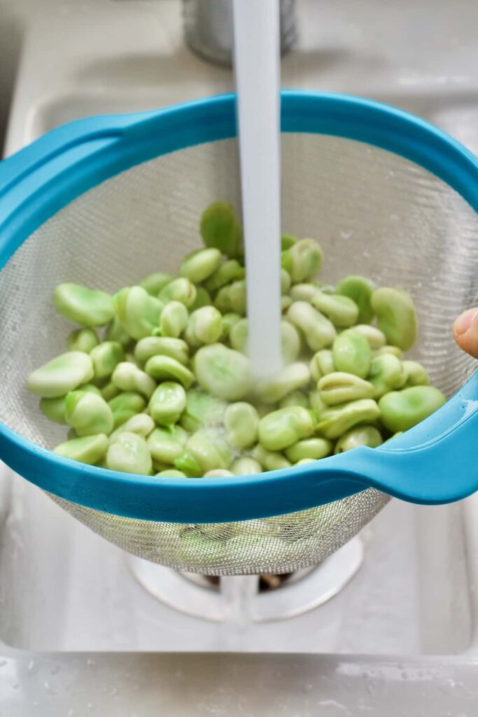 Cooked broad beans in a sieve being rinsed under the tap.