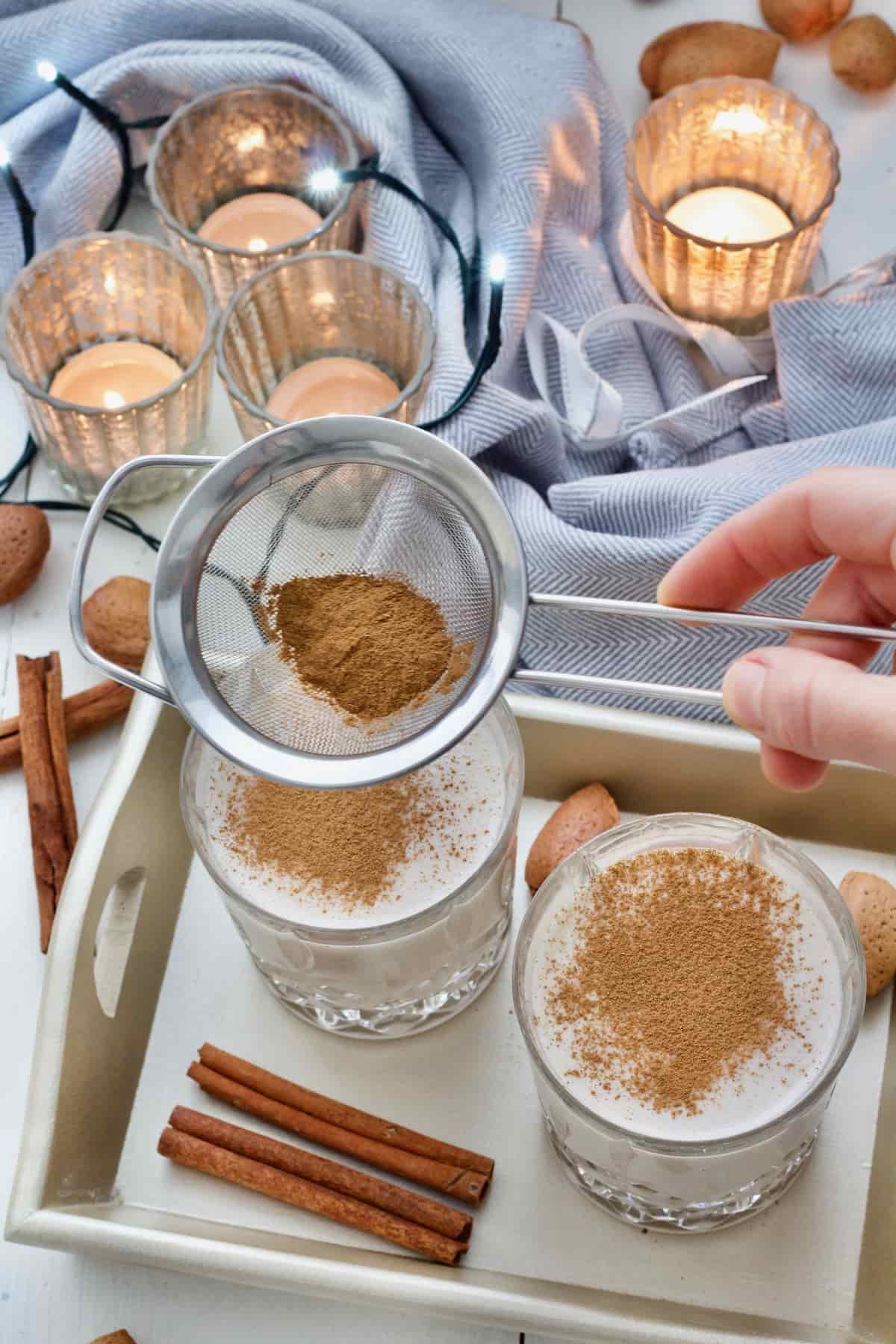 Hand holding small sieve with ground cinnamon in it over the glasses.