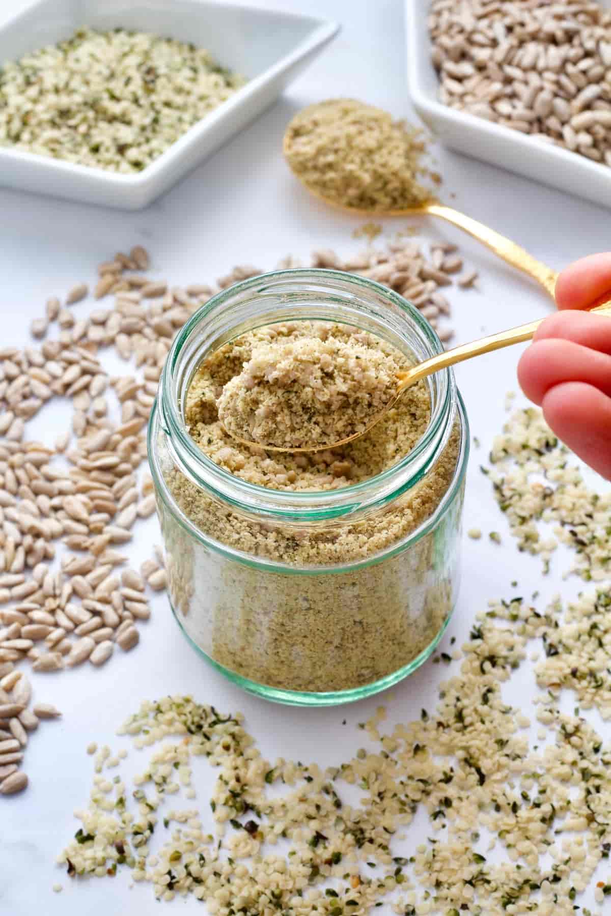 Spoon with vegan parmesan over the jar, scattered seeds around.