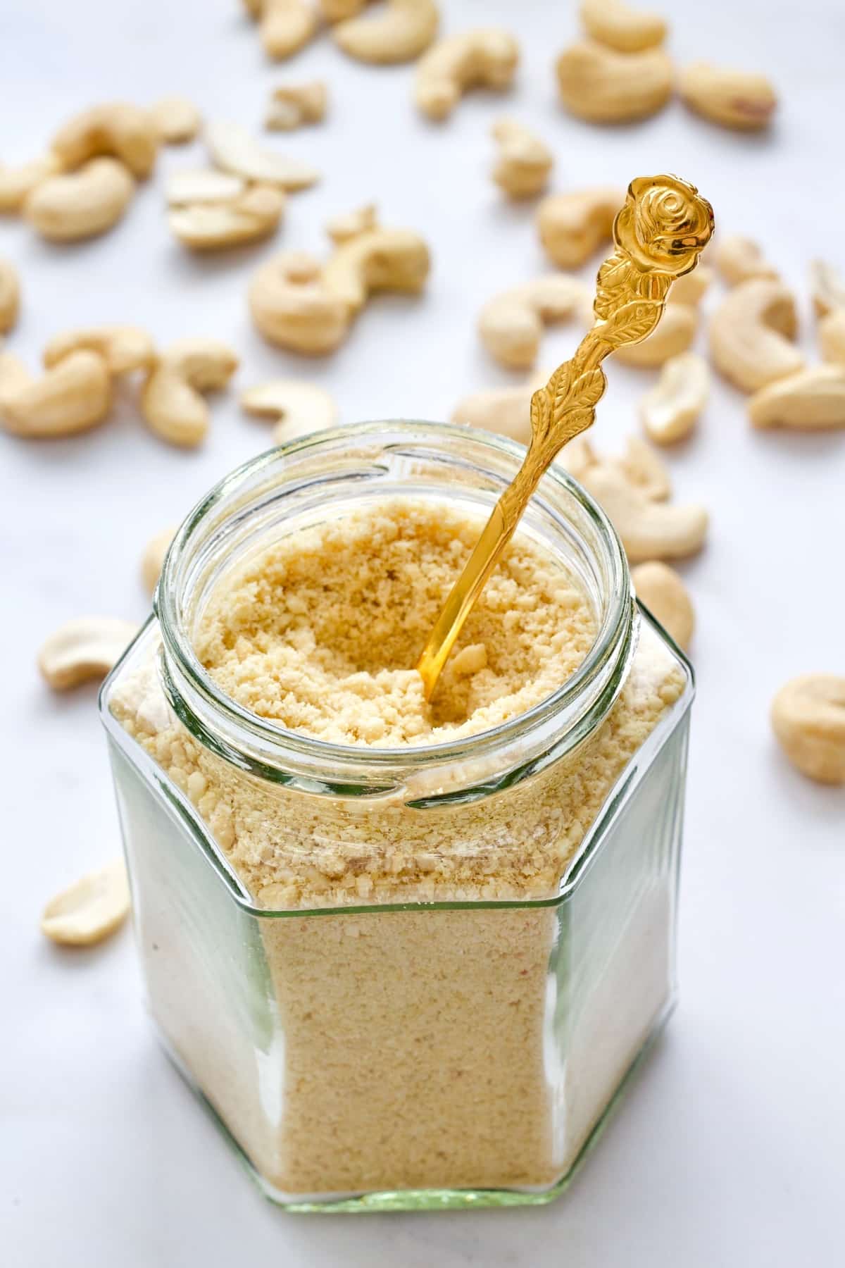 Vegan cashew parmesan in a jar with a spoon, scattered cashew nuts.