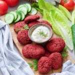 Beetroot falafel on a board with mayo dip in the middle, cucumber slices.