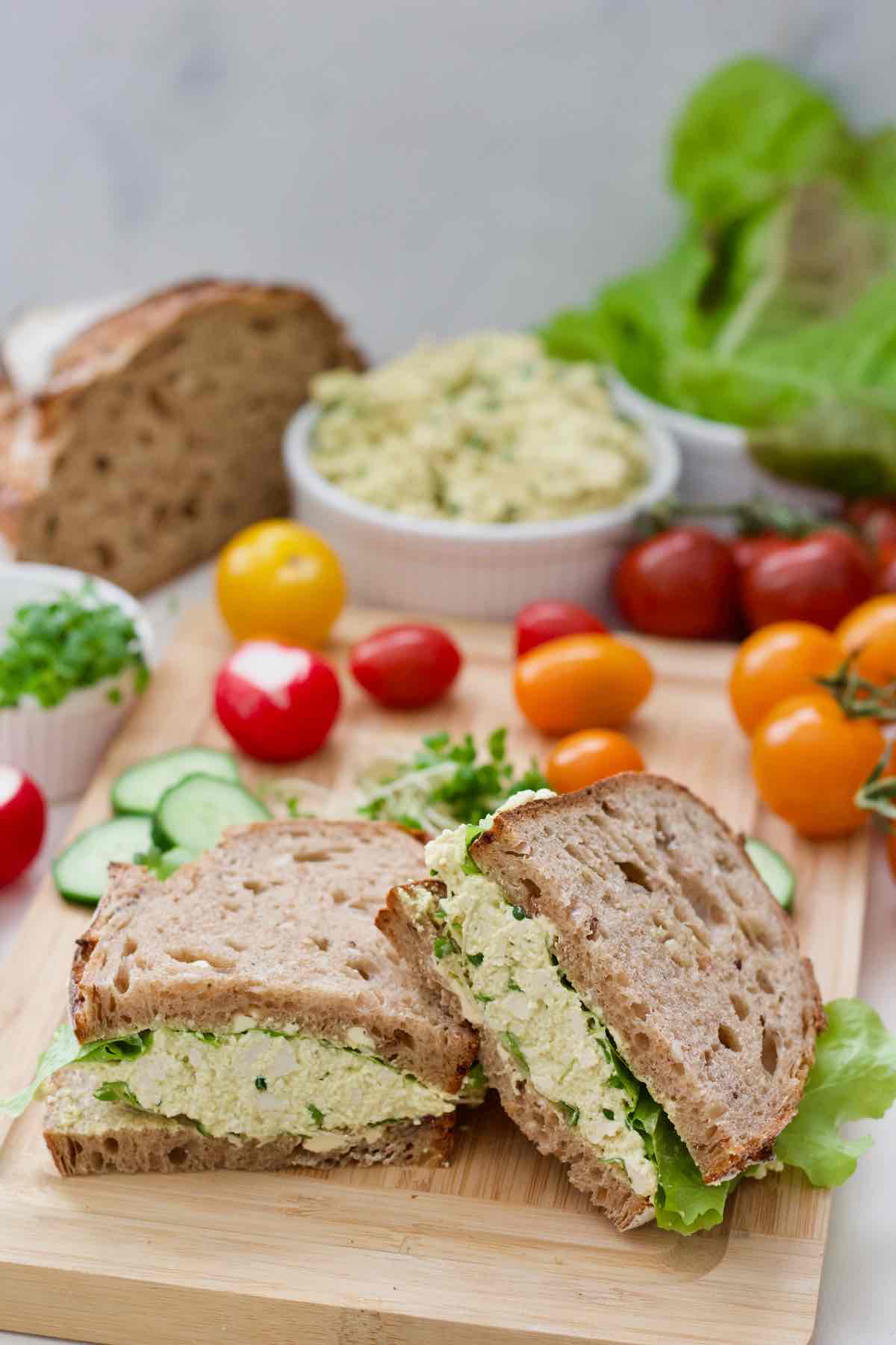 Two vegan egg salad sandwiches on the board with other veggie garnishes.