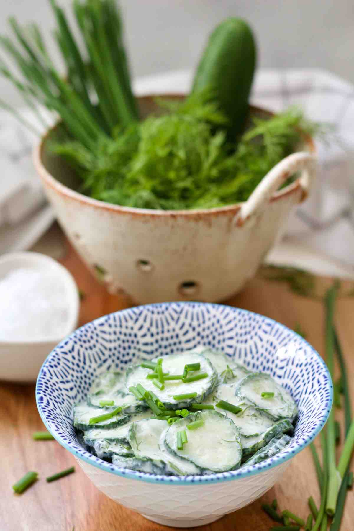 Mizeria in a bowl with green onions, dill and cucumber in a colander behind.