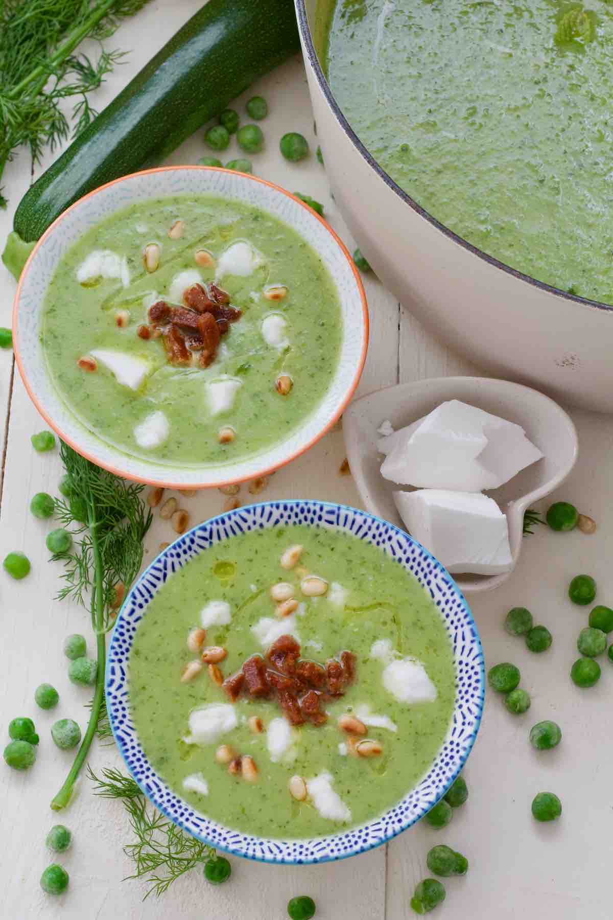 Two bowls of courgette soup with garnishes and peas scattered around.