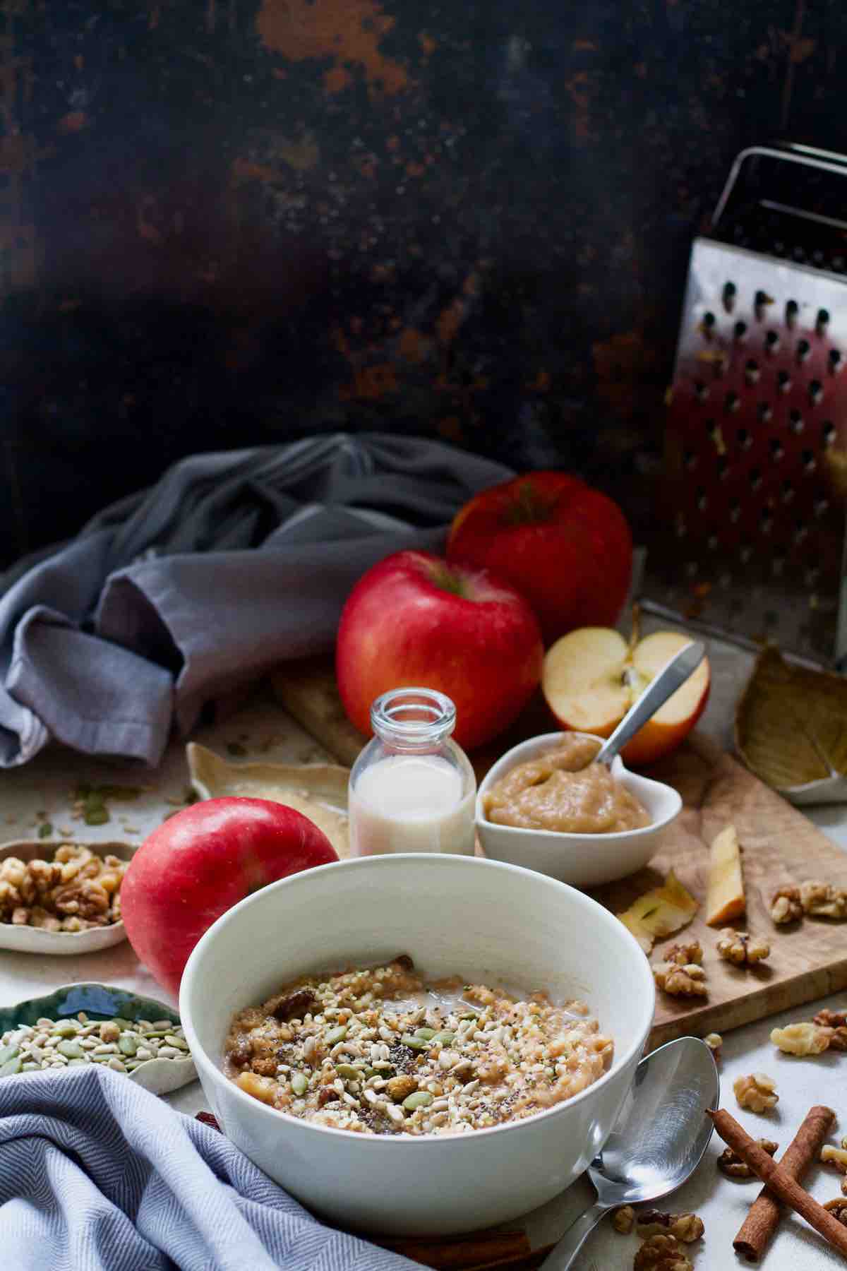 Breakfast setup with bowl of porridge, apples, plant milk, nuts, seeds and spices.