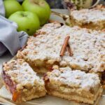 Square close up of szarlotka decorated with cinnamon sticks and with two slices cut.