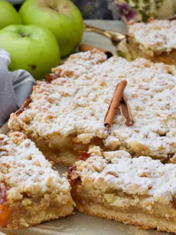 Square close up of szarlotka decorated with cinnamon sticks and with two slices cut.