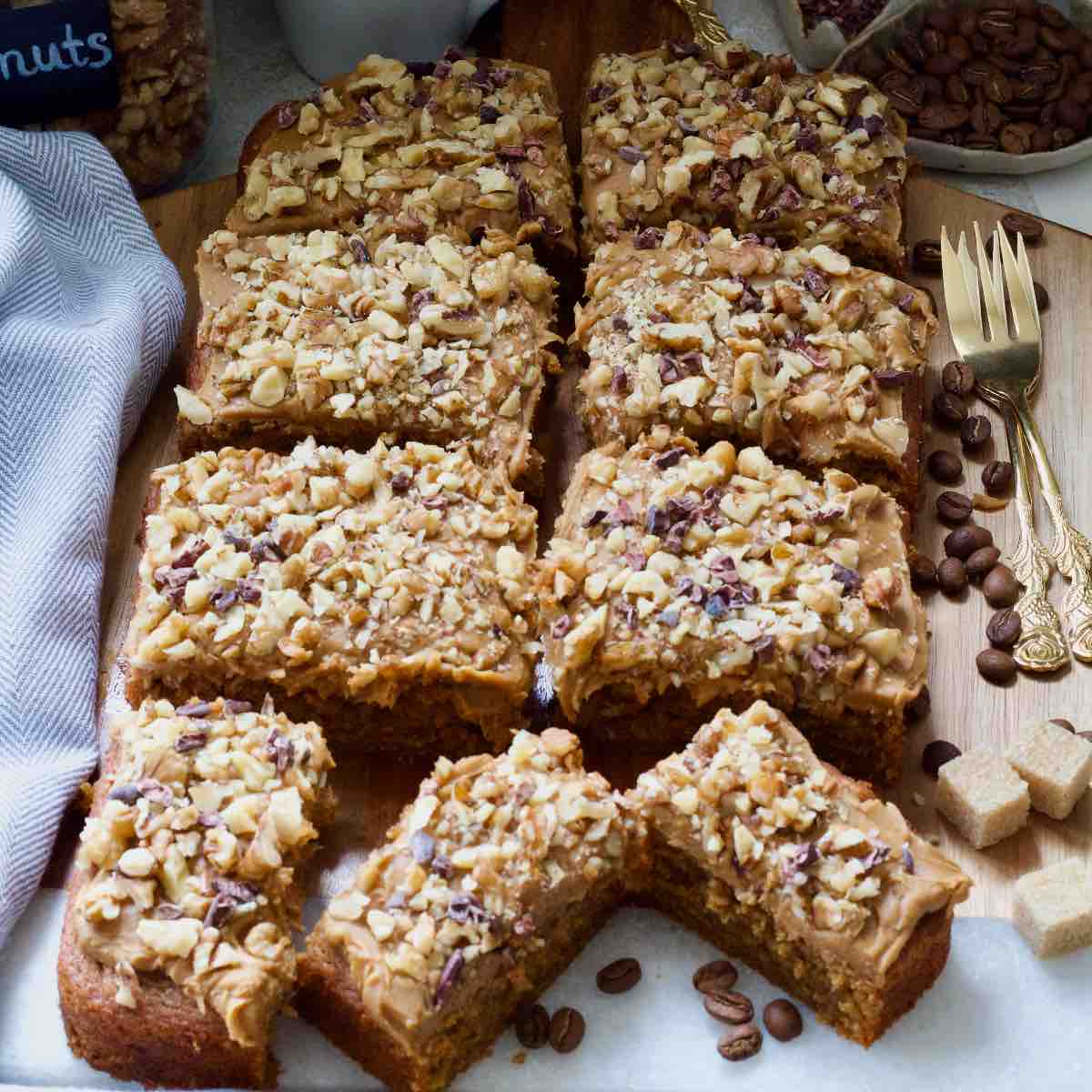 Slices of coffee and walnut cake arranged on a board, coffee beans, sugar cubes and forks.