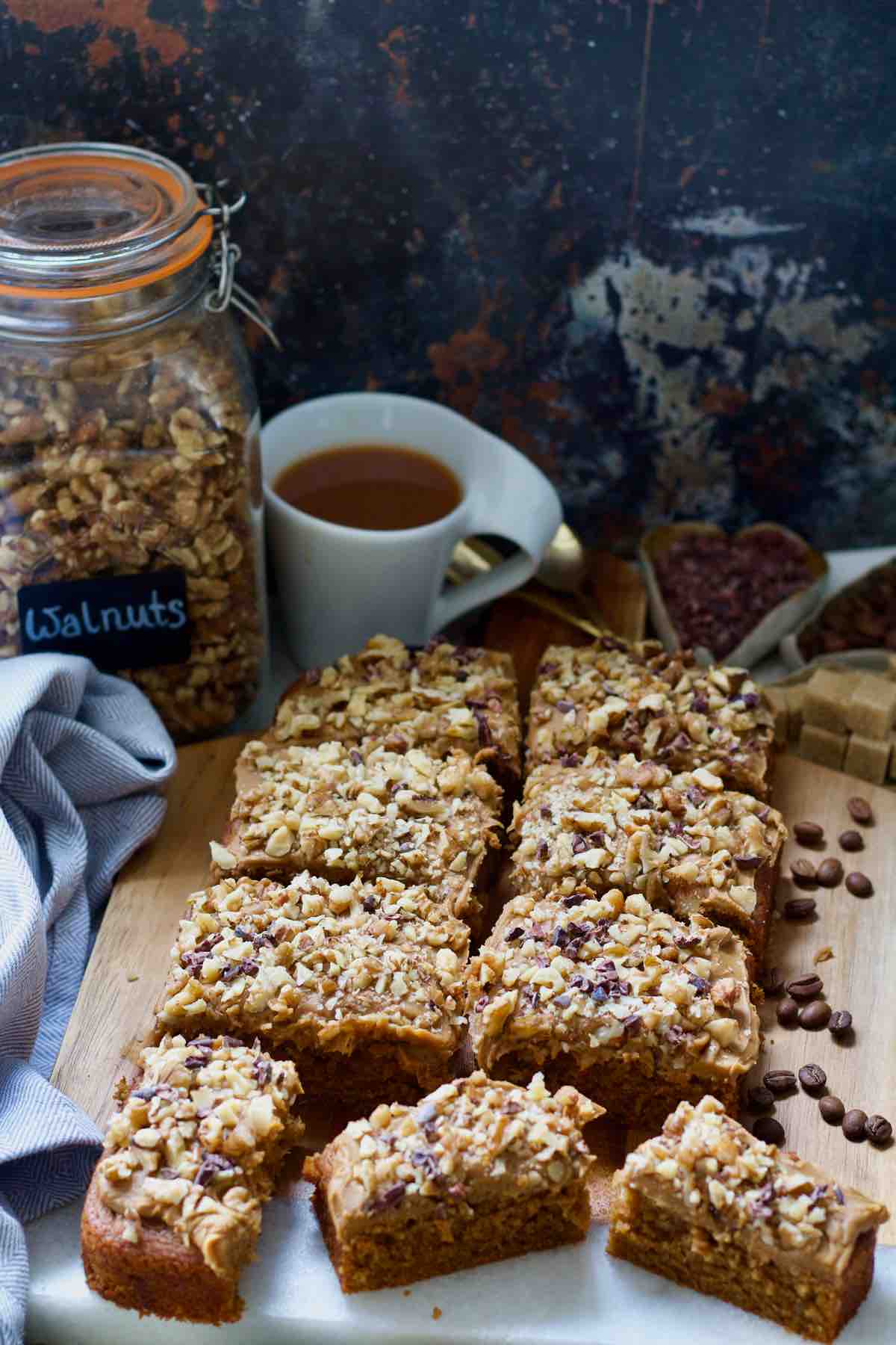 Coffee and walnut traybake portioned on the board with jar of walnuts and tea behind.