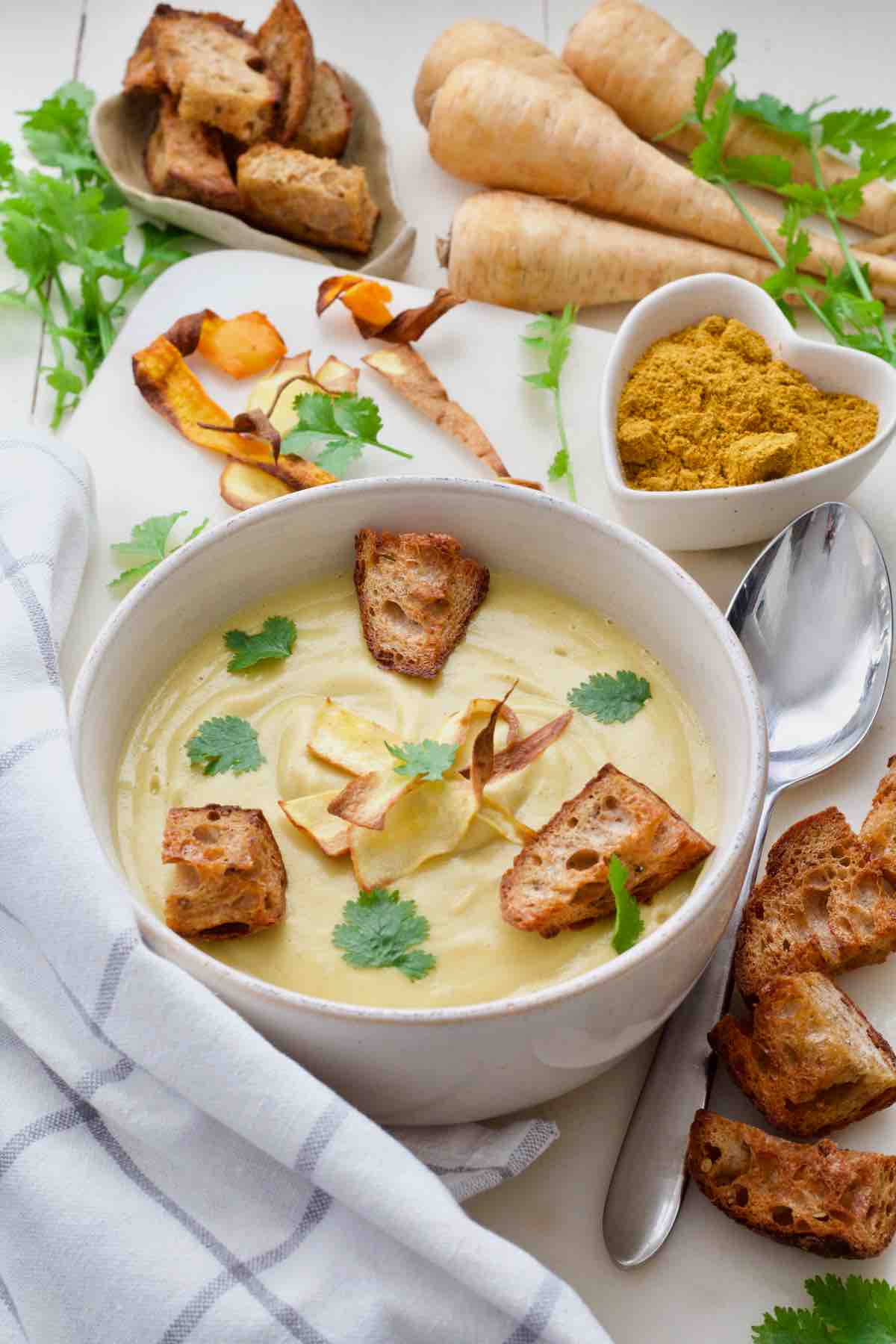 Bowl of soup surrounded by croutons, spices, veggie crisps, parsnips and herbs.