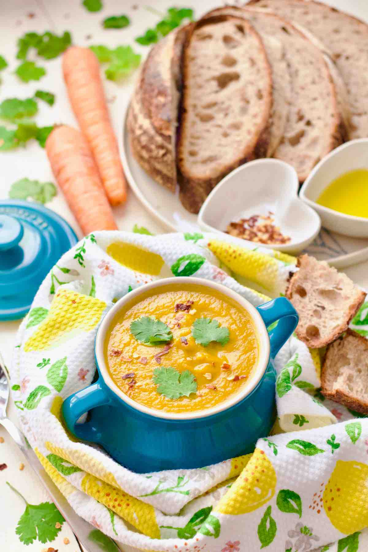 Carrot and coriander soup in serving bowl with bread, oil and chilli flakes.