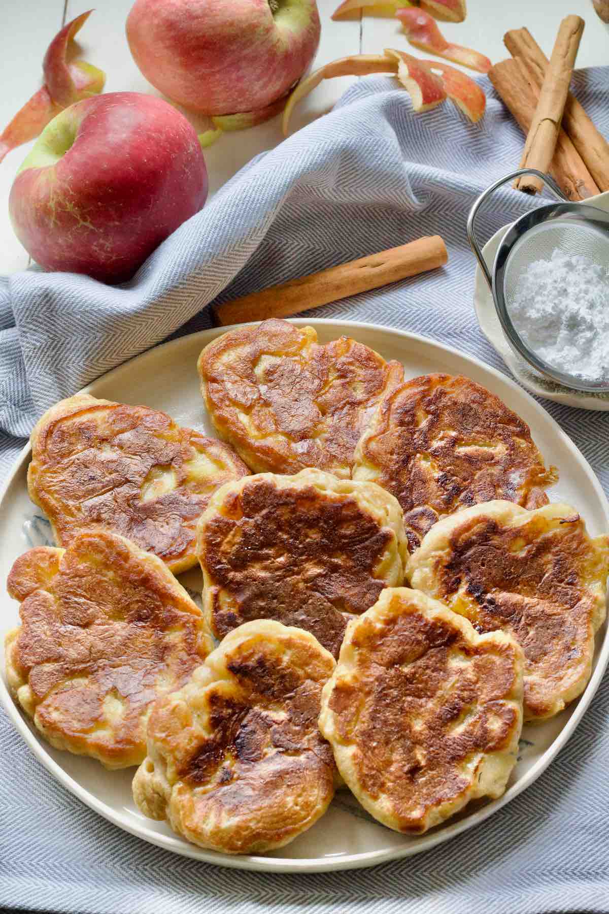 Round plate with apple pancakes, apples and cinnamon sticks behind it.