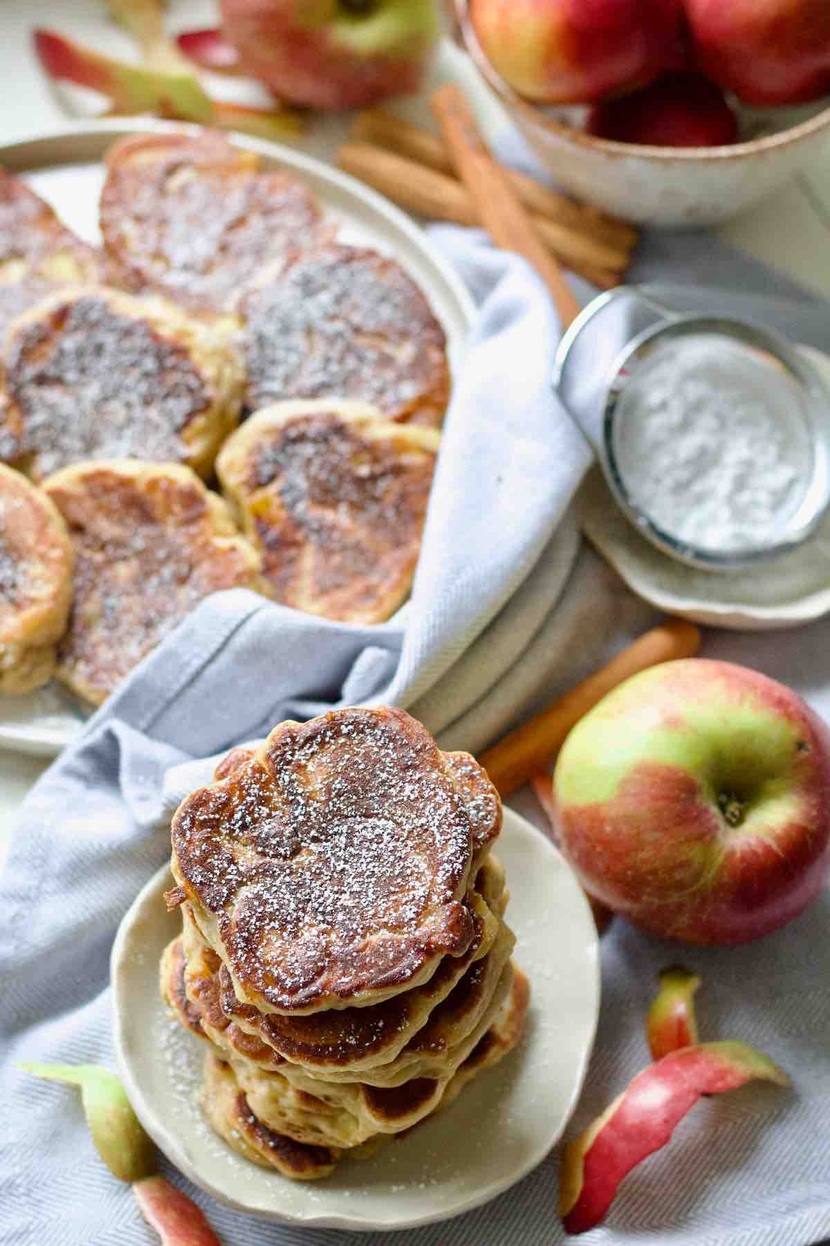 Stack of pancakes, an apple, sieve with icing sugar and plate with pancakes.