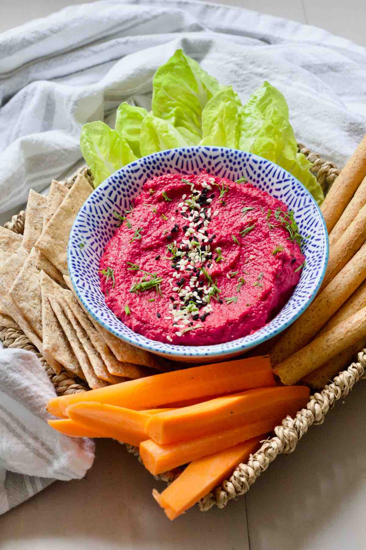 Bowl with hummus surrounded by salad leaves, carrot sticks, crackers and breadsticks.