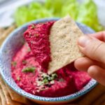 Hand holding a cracker dipped in pink hummus.