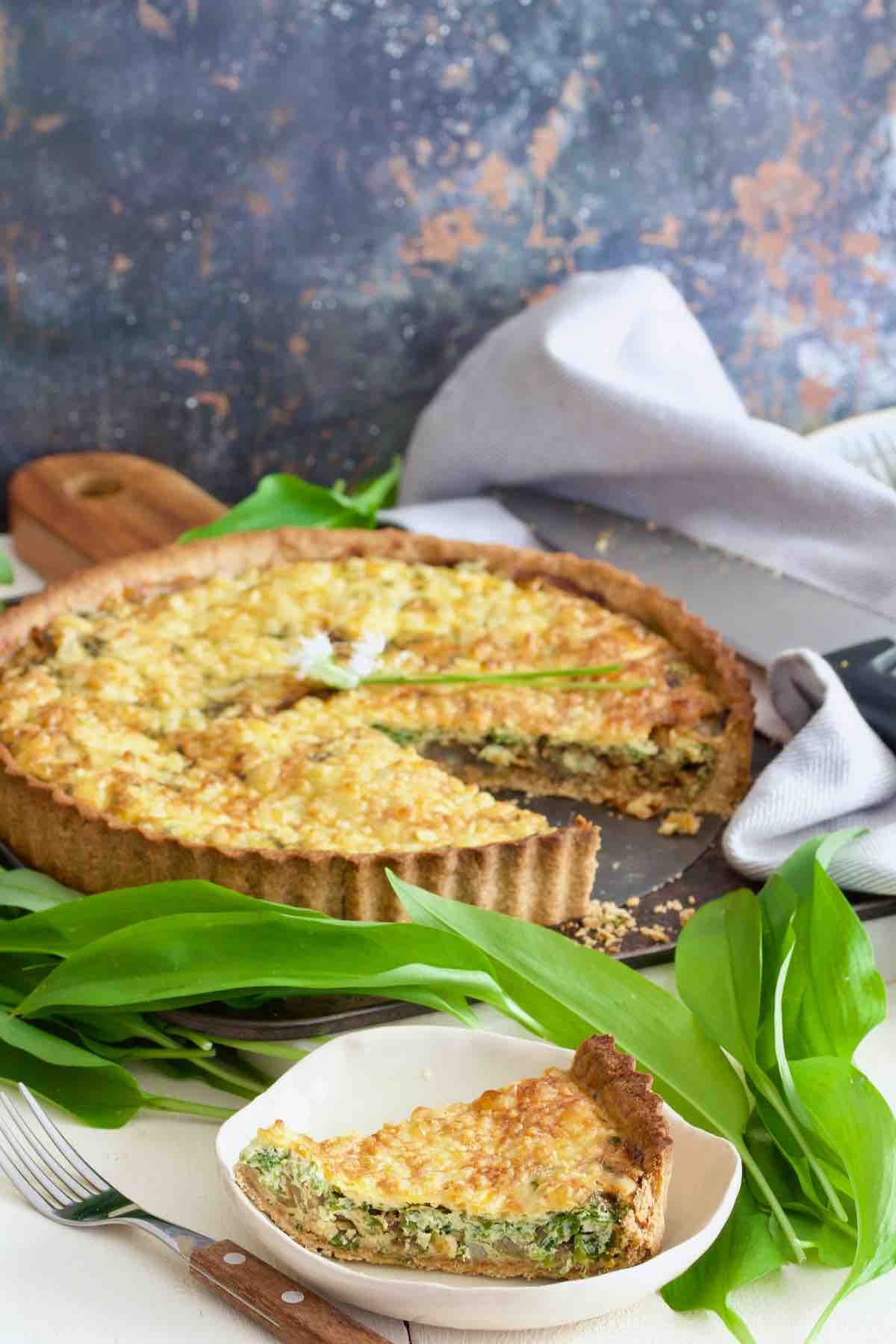 Cheese and onion quiche with single slice on a plate, wild garlic around.