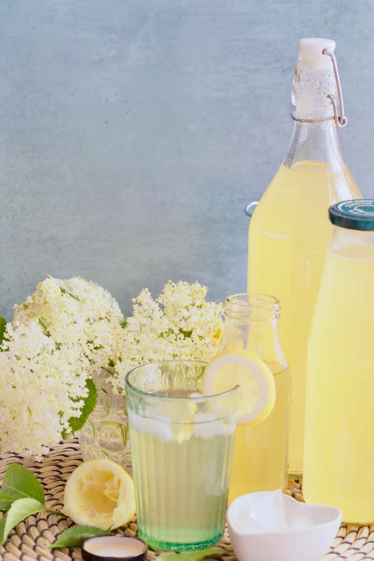 Small glass with diluted elderflower syrup with flowers behind and bottles of cordial.