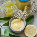 Small bowl with curd, open, jar, half a lemon and elderflowers and their leaves.