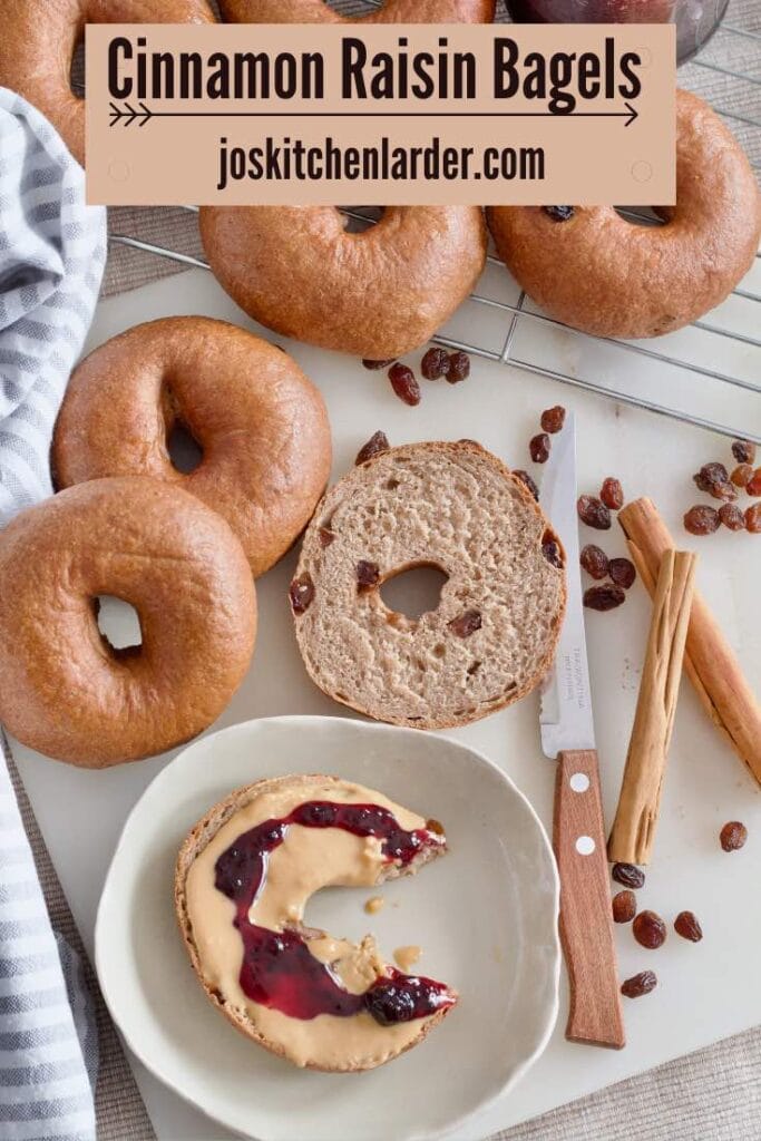 Bitten bagel with peanut butter and jam on a plate, more bagels, cinnamon and raisins.