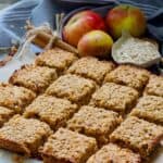 Neatly cut apple flapjacks with apples, bowl of oats and cinnamon sticks.