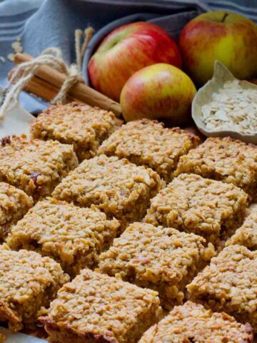 Neatly cut apple flapjacks with apples, bowl of oats and cinnamon sticks.
