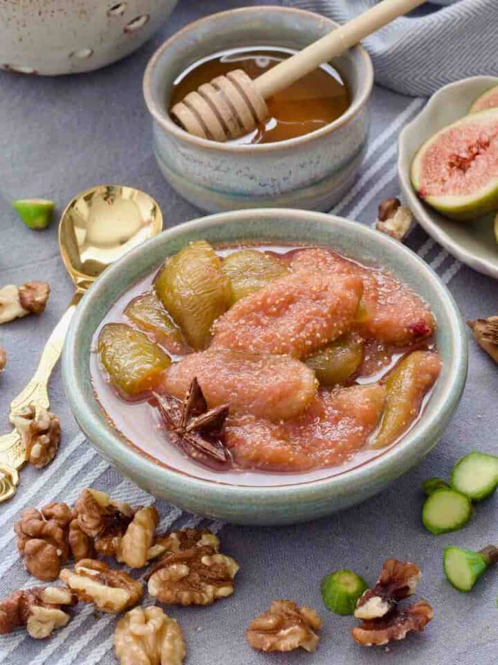Fig compote in a bowl with fig ends, walnuts, cinnamon sticks scattered around.