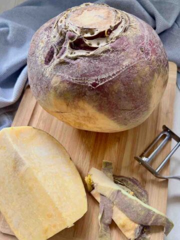 Whole swede on a cutting board with quarter swede chunk, peeler and some peelings.