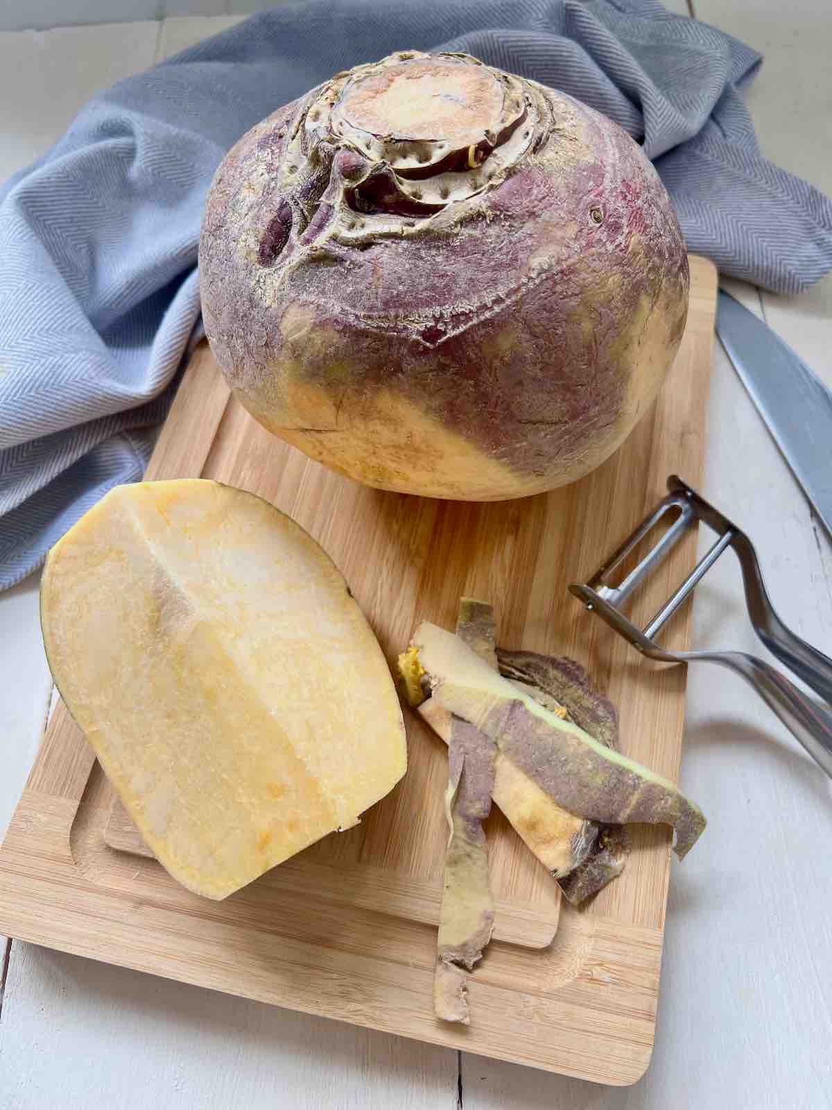 Whole swede on a cutting board with quarter swede chunk, peeler and some peelings.