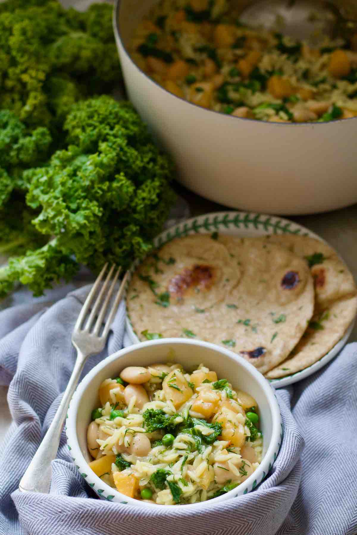 Portion of swede orzo in a bowl wit garlic flatbreads on a plate and pot with rest of orzo and some fresh kale behind.
