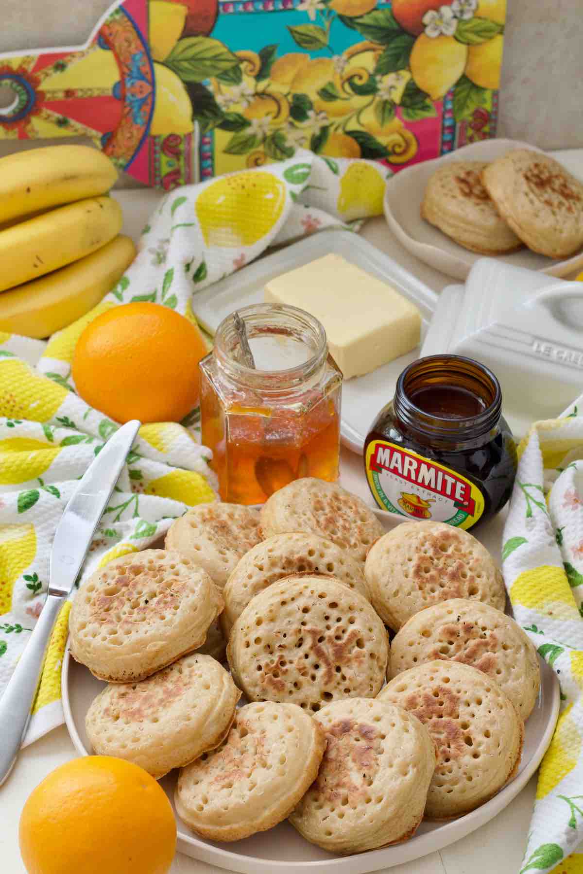 Plate with crumpets, marmite, jar with marmalade, butter, oranges and bananas.