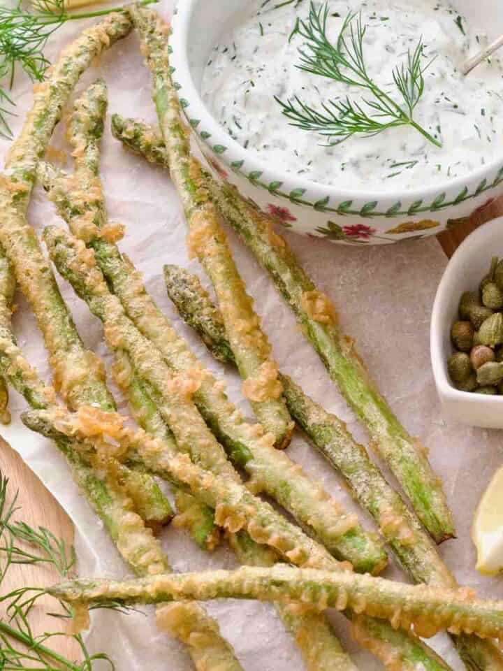 Square photo of asparagus tempura served with sauce and lemon wedges.