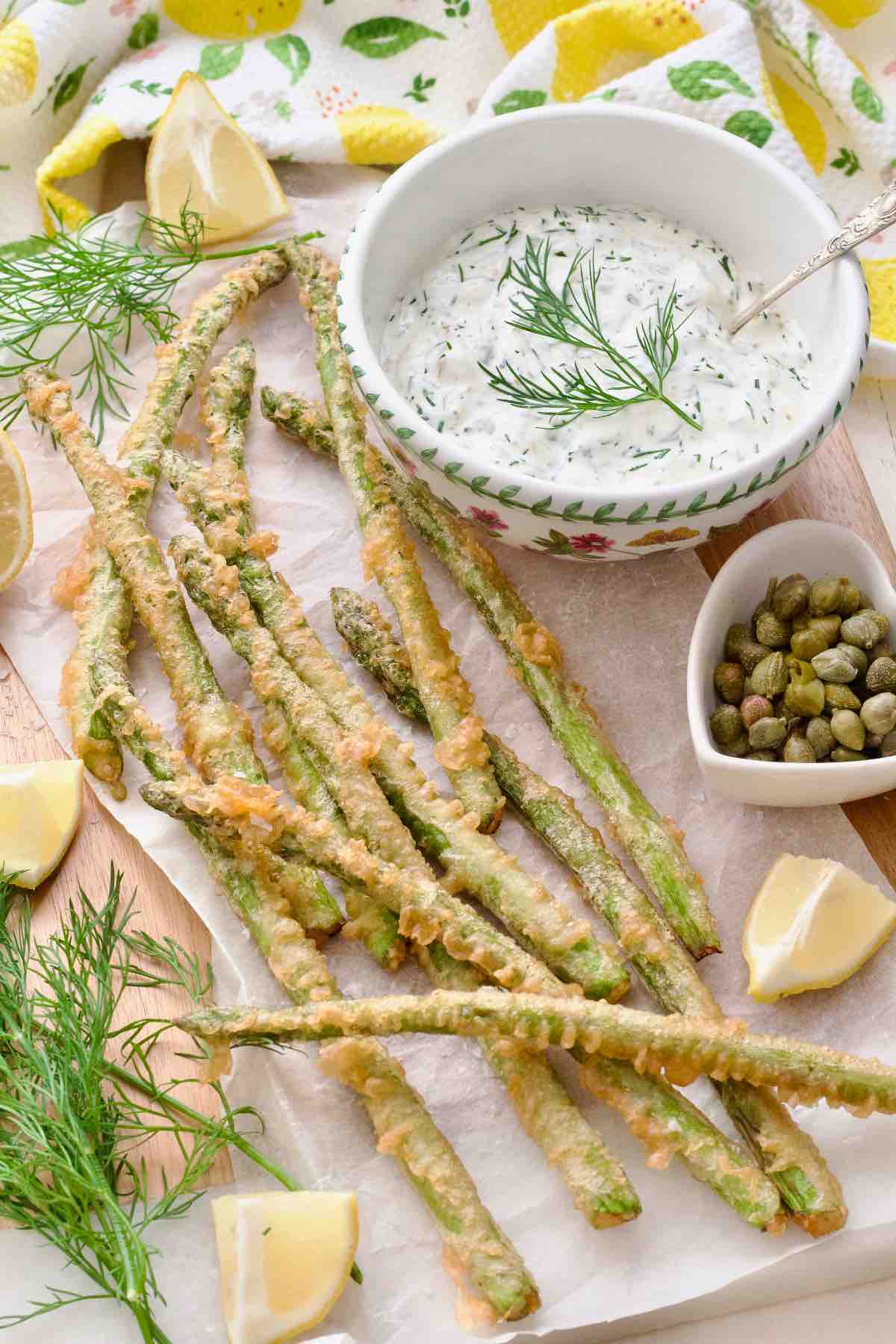 Asparagus tempura presented on a board covered with paper with tartar sauce and lemon wedges.