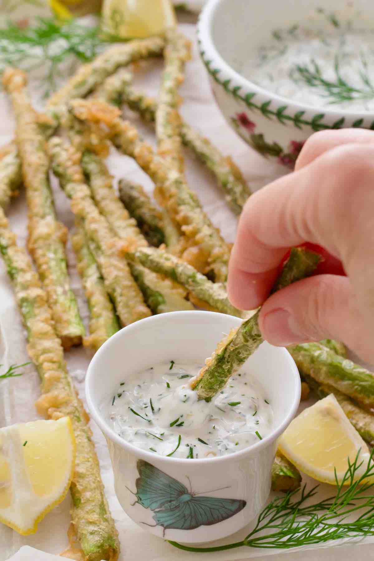 Hand dipping asparagus tempura in a small bowl with tartar sauce.