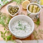 Square image of tartar sauce in a serving bowl, gherkins, lemon and capers behind it.