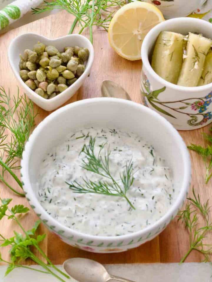 Square image of tartar sauce in a serving bowl, gherkins, lemon and capers behind it.