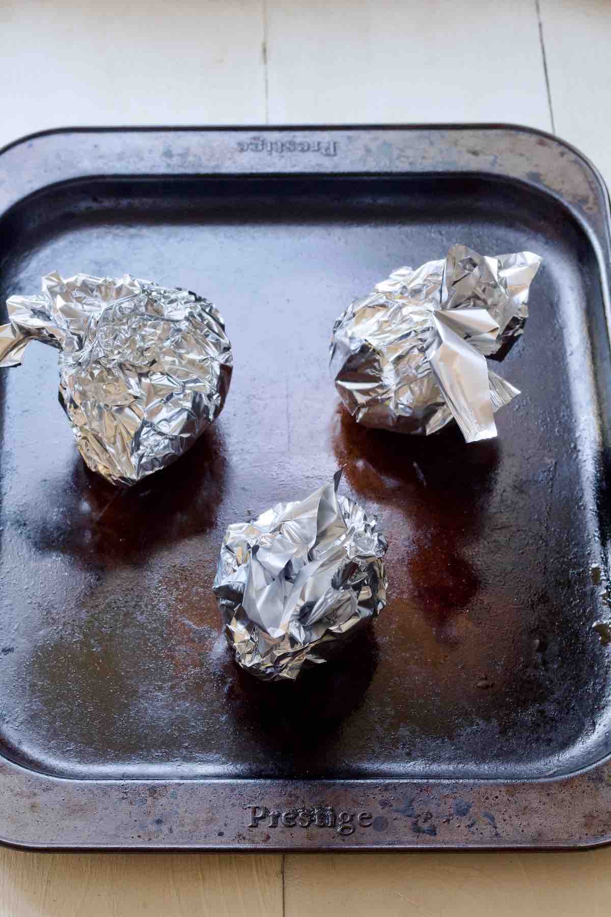 Three beets wrapped in aluminium foil on a baking tray ready for roasting.
