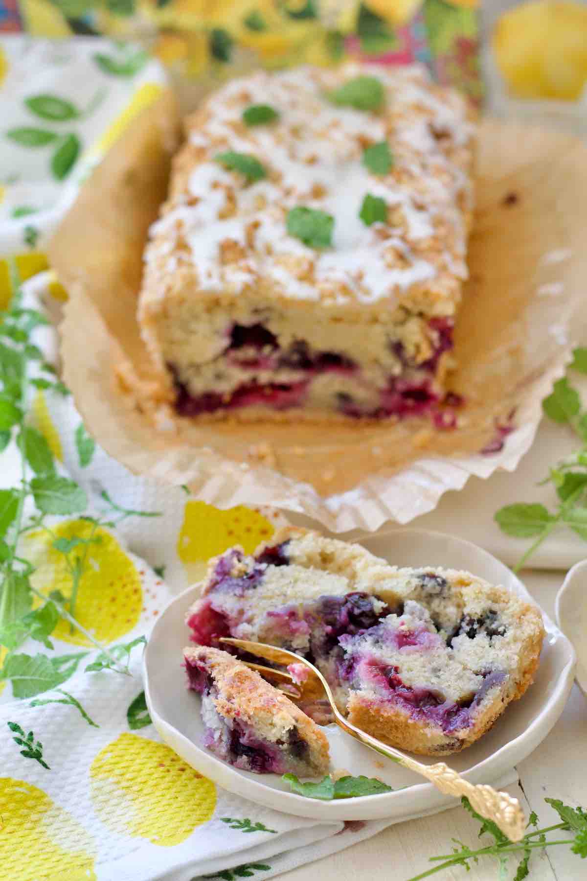 Slice of lemon and blueberry loaf cake on a plate and rest of the loaf behind.