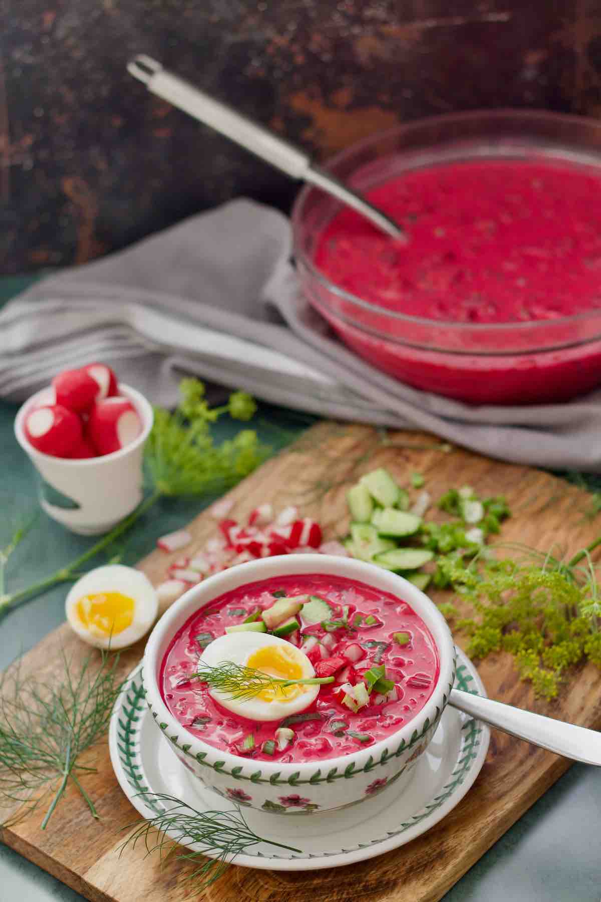 Polish cold beet soup served in a bowl with accompaniments around it and large bowl of chłodnik in the background.