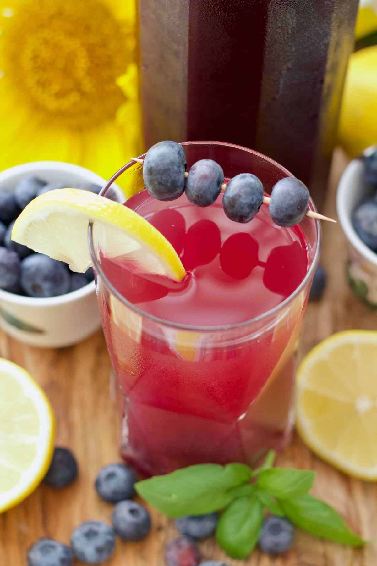 Diluted blueberry cordial in a glass with blueberries on a stick and a slice of lemon.