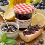 Close up of labelled blueberry jam jar, jam in a bowl and a piece of jam topped baguette.